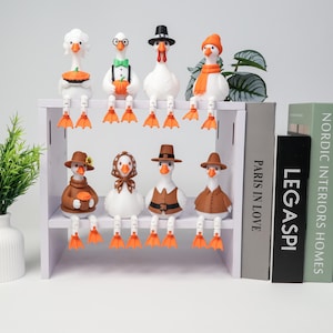 Könnte beinhalten: Acht weiße Gänsefiguren mit orangefarbenen Füßen und baumelnden Beinen auf einem zweistöckigen weißen Regal. Die Gänse tragen verschiedene saisonale Accessoires wie Hüte, Schals und Outfits, einige halten kleine Gegenstände wie Kürbisse und Kuchen.
