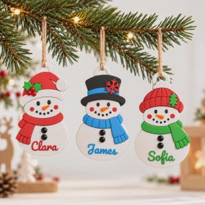 Könnte beinhalten: Drei weiße Schneemann-Anhänger hängen an einem Tannenzweig. Der linke Schneemann trägt eine rote Mütze und einen roten Schal mit dem Namen Clara. Der mittlere Schneemann trägt einen schwarzen Zylinder und einen blauen Schal mit dem Namen James. Der rechte Schneemann trägt eine rote Mütze und einen grünen Schal mit dem Namen Sofia.
