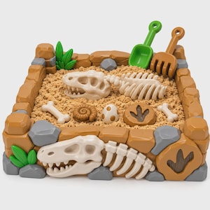 May include: A square sandbox playset featuring a tan sand-filled basin with brown stone-like borders, green plastic plants, and gray rocks. Inside are white dinosaur skeleton pieces, a brown ammonite, a spotted egg, and a footprint fossil.