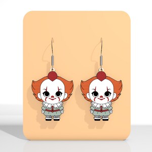 May include: A pair of dangle earrings featuring a cartoon clown design. The earrings have a red heart-shaped top, orange hair, white face paint, and a gray outfit. The earrings are displayed on a peach-colored background.