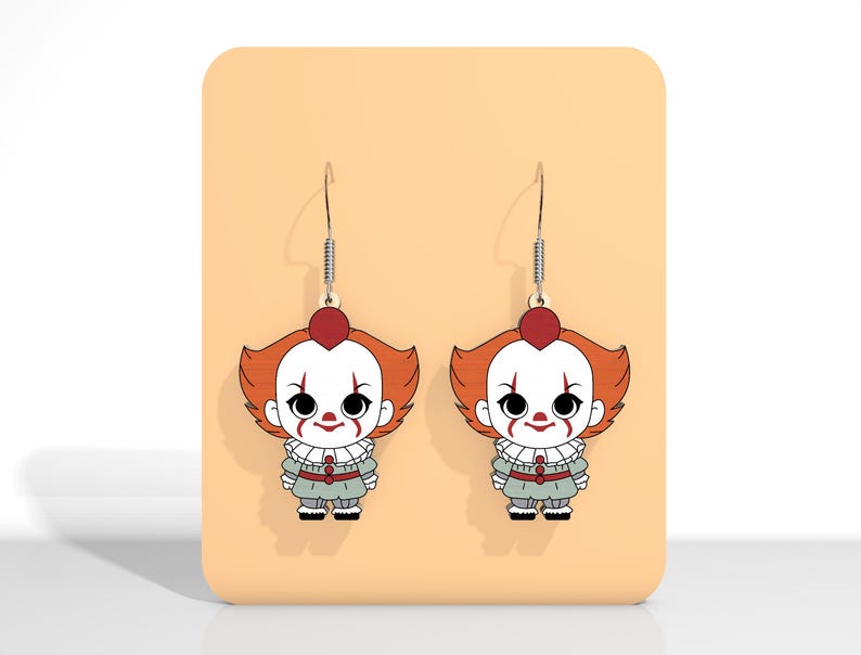 May include: A pair of dangle earrings featuring a cartoon clown design. The earrings have a red heart-shaped top, orange hair, white face paint, and a gray outfit. The earrings are displayed on a peach-colored background.