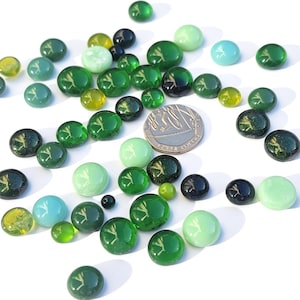 May include: A collection of glass marbles in various shades of green, yellow, and blue, scattered on a white surface. The marbles vary in size, with a few dark green and black marbles. A coin is also present, adding scale to the image.