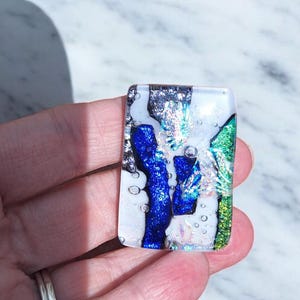 May include: Rectangular glass pendant with a dichroic design. The pendant features a mix of colors, including blue, green, silver, and white. The glass has a textured appearance with bubbles and a glossy finish. The pendant is held in a hand.