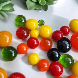 May include: A selection of colourful glass marbles in various sizes and shades. The marbles are in shades of yellow, red, orange, green, and black. The marbles are scattered on a white surface, with a green plant in the background.