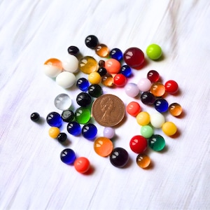 May include: A selection of multi-coloured glass marbles of varying sizes, including red, blue, green, yellow, and orange. A copper coin is included for size reference. The marbles are arranged on a white, textured fabric.