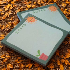 Peut inclure: Deux blocs-notes carrés avec un fond bleu clair et une bordure verte. Chaque bloc-notes porte le mot "notes" imprimé en vert et une illustration de soleil souriant. Un petit pot de fleurs rose avec une plante verte est dans le coin.