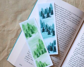 May include: Two handmade watercolor bookmarks with forest scenes. One bookmark features a green forest, the other a blue forest. The bookmarks are rectangular and placed on an open book.