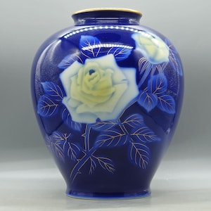 May include: A cobalt blue vase featuring a large, pale yellow rose and a smaller rosebud. The floral design is accented with delicate gold outlines on the petals and leaves. The vase has a rounded body, a narrow neck, and a thin gold rim at the top.