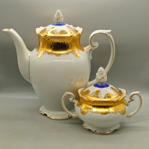 Puede incluir: Un juego de té de porcelana blanca con adornos y detalles dorados. El juego incluye una tetera y un azucarero. La tetera tiene un asa y un pico curvos. El azucarero tiene dos asas. Ambas piezas tienen detalles dorados ornamentados.