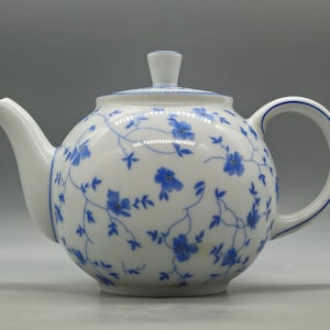 Op de afbeelding: Een witte porseleinen theepot met een delicaat blauw bloemenrankpatroon. De theepot heeft een rond lichaam, een gebogen tuit, een C-vormig handvat en een bijpassend deksel met een kleine knop. Dunne blauwe lijnen accentueren de rand en het handvat.