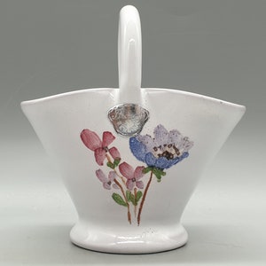 May include: White ceramic basket with a handle, featuring a floral design of pink and purple flowers with green stems. The basket has a silver metal tag with the word "Bellevue" on it.