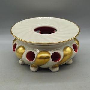 Pode incluir: Um aquecedor redondo de cerâmica com topo branco, borda dourada e padrão em espiral. A base branca apresenta detalhes dourados em forma de gota e aberturas circulares que revelam um interior vermelho vibrante. Apoia-se em pequenos pés.
