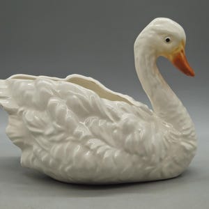 Pode incluir: Um vaso ou taça decorativa de cerâmica branca em forma de cisne, com acabamento brilhante, detalhes de penas moldadas no corpo e um pescoço liso com um bico pintado de laranja. O cisne está representado em posição de repouso com cavidade.