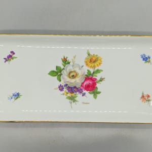 Könnte beinhalten: Ein rechteckiges Tablett aus weißem Porzellan mit abgerundeten Ecken und einem goldfarbenen Rand. In der Mitte befindet sich ein bunter Blumenstrauß aus weißen, gelben, rosa und violetten Blüten, ergänzt durch kleine Blumenmotive in den Ecken.