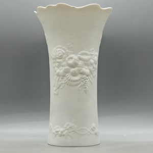 May include: A tall, white ceramic vase with a flared top and a detailed floral relief design. The vase features embossed roses, other flowers, and foliage. The vase is approximately 30 cm tall.