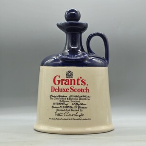 Puede incluir: Una jarra de cerámica con la parte superior y el asa de color azul oscuro y una base de color crema. El texto "Grant's Deluxe Scotch" está impreso en rojo. La jarra tiene forma redondeada y un tapón.