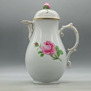 Puede incluir: Una cafetera de porcelana blanca con un motivo de rosa rosa pintado a mano con hojas verdes en el cuerpo. La tapa está rematada con un remate esculpido en forma de capullo de rosa rosa. El asa, el pico y el borde tienen detalles en oro.