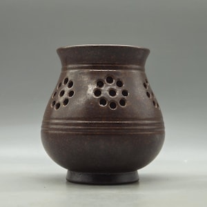 May include: A brown ceramic vase with a rounded body and a flared rim. The vase features decorative circular cutouts and horizontal grooves. The vase is approximately 13 cm tall and has a dark brown colour.
