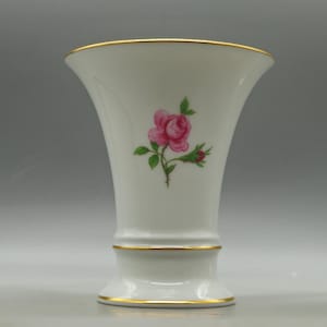 May include: White porcelain vase with a flared top and a gold rim. A pink rose with green leaves and buds is painted on the front. The base also has a gold trim. A decorative floral vase.
