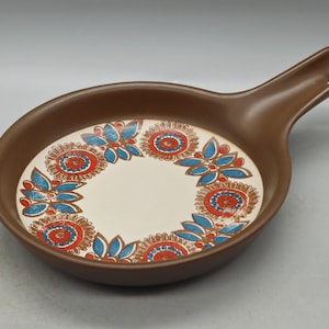 May include: A brown ceramic serving dish with a handle. The dish is decorated with a white background and a colorful floral pattern in shades of red, blue, and orange.