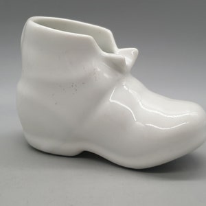 Peut inclure: Un vase en céramique blanche en forme de chaussure. Le vase est verni et a une finition lisse et brillante.