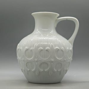 May include: White ceramic pitcher with a handle and a decorative embossed pattern. The pitcher has a bulbous body and a narrow neck. The embossed design features vertical lines and repeating floral-like shapes. The pitcher is a classic piece for serving beverages.