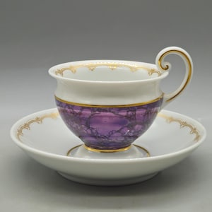 May include: White teacup and saucer set with gold trim. The cup features a purple marbled design around the middle. The handle and rim of the cup and saucer are accented with gold detailing. The saucer is round.