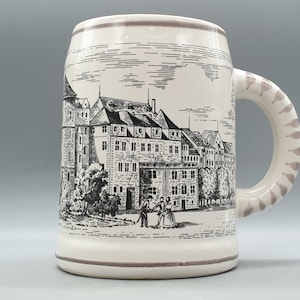 Könnte beinhalten: Ein weißer Keramik-Bierkrug mit einer Schwarzweiß-Illustration eines Gebäudes und von Menschen. Der Henkel ist mit einem braunen Muster verziert.
