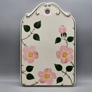 May include: White ceramic cutting board with a floral design. The board features pink roses with yellow centers and green leaves on a white background. The board has a decorative handle with a hole in the center.
