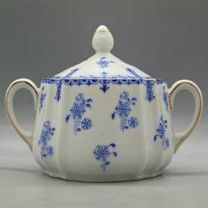 May include: A white porcelain sugar bowl with a lid, adorned with blue floral patterns and gold accents. The bowl has two handles and a decorative finial on the lid. A classic piece for serving sugar.