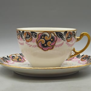 Puede incluir: Juego de taza y platillo de té color crema con un diseño floral en negro, rosa y dorado. La taza tiene un asa y borde dorados, y el platillo tiene un borde dorado a juego. El juego es probablemente vintage o antiguo.