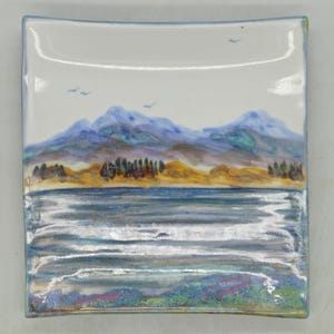 Pode incluir: Prato quadrado de cerâmica com uma paisagem pintada à mão. A obra retrata um lago com montanhas ao fundo, em tons de azul, verde e castanho. O prato tem um fundo branco e uma borda azul.