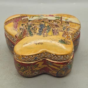 Puede incluir: Caja de baratijas de cerámica en forma de corazón con una tapa que presenta una escena detallada de personas con vestimenta tradicional japonesa. La caja tiene una base de color crema con detalles en rojo y dorado, de unos 15 cm de ancho.