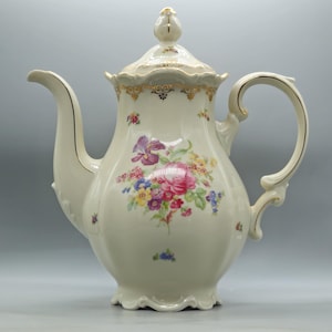 Può includere: Caffettiera in porcellana color crema con un motivo floreale. La caffettiera presenta un beccuccio curvo, un manico ad anello e un coperchio con un pomello decorativo. Rifiniture dorate accentuano i bordi e il manico.