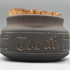 Puede incluir: Un tarro de tabaco de cerámica marrón con tapa de corcho. El tarro tiene un diseño decorativo con la palabra "Tobak" escrita en una fuente estilizada.