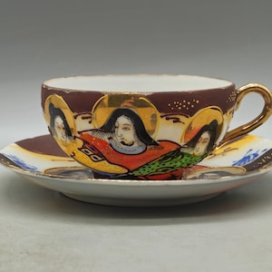 Puede incluir: Juego de taza y platillo de té vintage. La taza y el platillo son blancos con un borde marrón. La taza presenta tres figuras pintadas con halos y detalles dorados. El asa y el borde son dorados. El platillo tiene un diseño a juego.