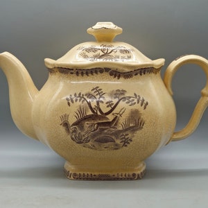 May include: A cream-colored teapot with a brown floral and animal design. The teapot has a lid with a knob on top and a handle on the side.