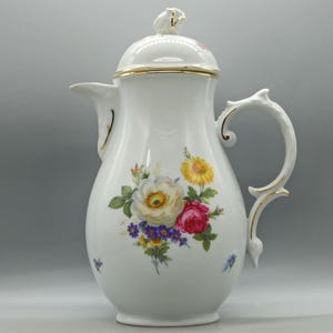 May include: White porcelain coffee pot with a lid, gold trim, and a floral design featuring yellow, red, and white roses. The handle and spout are elegantly shaped. A decorative piece for serving coffee or tea.