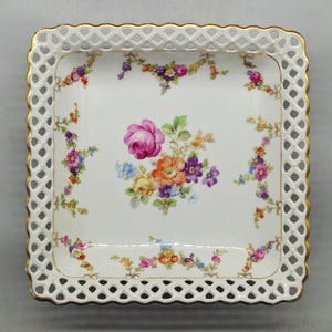 May include: Square, white porcelain tray with a floral design. The tray has a pierced, openwork border with a gold trim. The center of the tray features a bouquet of pink, orange, purple, and yellow flowers. Floral sprigs decorate the corners.