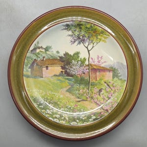 Puede incluir: Un plato redondo de cerámica con un borde verde y un centro marrón. El plato presenta una escena pintada de un paisaje campestre con una casa, árboles y flores.