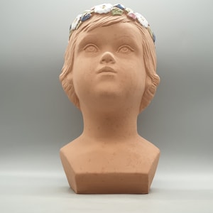 Puede incluir: Busto de terracota de un niño con una corona de flores. La corona está hecha de flores blancas, azules y verdes.