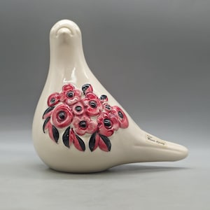 Puede incluir: Figurita de paloma de cerámica color crema con un grupo de acentos florales rojos y negros en su costado. El ave tiene un acabado liso y brillante y un pico largo y puntiagudo. La figurita es un artículo decorativo.