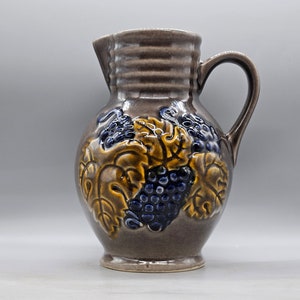 May include: A brown ceramic pitcher with a handle and a design of grapes and leaves in shades of blue, yellow, and brown.