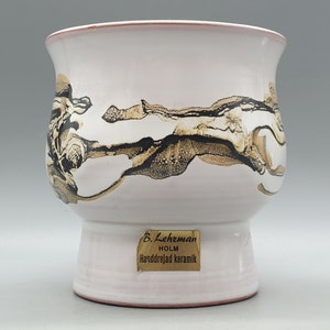 Könnte beinhalten: Weiße Keramikvase mit einem abstrakten Design in Schwarz und Gold. Die Vase hat eine breite Öffnung und einen runden Boden. Am Boden der Vase befindet sich ein goldenes Etikett mit der Aufschrift "B. Lehzman Holm Handdrejad Keramik".