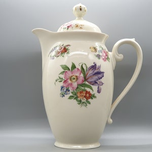 Puede incluir: Una cafetera de cerámica blanca con un diseño floral. La cafetera tiene una tapa con un pomo dorado y un asa en el lateral. Las flores son rosas, moradas, azules y amarillas.