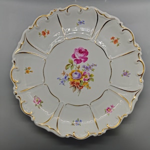 Puede incluir: Un plato de porcelana blanca con un borde dorado y un diseño floral. El plato tiene ocho secciones y un motivo floral central con una rosa rosa y flores amarillas, naranjas y moradas.