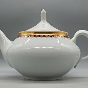 Könnte beinhalten: Weiße Porzellan-Teekanne mit Deckel und Henkel. Die Teekanne hat ein gold-rotes Zierband um den Rand und den Deckel. Der Ausguss ist gebogen und der Griff ist eine Schlaufe. Die Teekanne ist zum Servieren von Tee bestimmt.