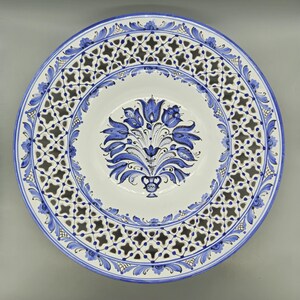 Op de afbeelding: Een witte decoratieve keramische plaat met een blauw bloemmotief. De plaat heeft een centraal boeket van blauwe bloemen en een geperforeerde rand met een zich herhalend geometrisch patroon. De rand is versierd met blauwe bloemaccenten.