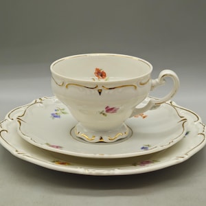 May include: A white porcelain teacup and saucer set with a matching plate, featuring delicate hand-painted floral motifs in red, pink, and blue. The edges of the cup, saucer, and plate are decorated with gold trim and scalloped detailing.