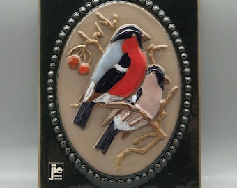 May include: A ceramic tile with a green background and a raised oval design. The oval features two birds perched on a branch with red berries. The birds are painted in shades of red, black, white, and gray. The tile is signed "JIE GANTKUTA SWEDEN".